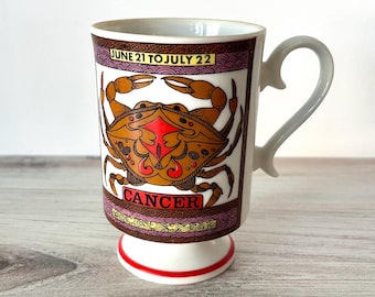 May include: A white ceramic mug with a crab zodiac design. The mug features a brown crab illustration with red accents, the word "CANCER", and the dates "JUNE 21 TO JULY 22". The mug has a unique handle and a red stripe at the base.