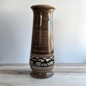May include: Tall, brown ceramic vase with a flared top and a decorative band. The vase features a base and a patterned design with white, green, and brown elements. The vase has a height of approximately 12 inches.