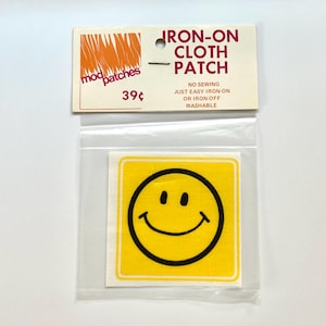 May include: A square yellow cloth patch featuring a black smiley face graphic, enclosed in a clear plastic bag with a cardboard header. The header reads IRON-ON CLOTH PATCH, mod patches, 39 cents, NO SEWING, JUST EASY IRON-ON OR IRON-OFF, WASHABLE.