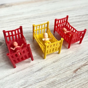 Könnte beinhalten: Drei Miniatur-Kinderbetten in Rot und Gelb, jedes mit einer kleinen Babyfigur. Die Betten haben ein klassisches Design mit vertikalen Stäben. Die Babys tragen Windeln. Das Bild ist aus einem leicht erhöhten Winkel aufgenommen.