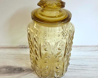 May include: A vintage amber glass jar with a lid. The jar has a square shape with embossed floral patterns. The lid is round with a flower-shaped knob. The jar is made of transparent glass, allowing the contents to be visible.
