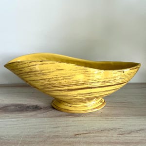 May include: A yellow ceramic bowl with an elongated, asymmetrical boat-like shape and a pedestal base. The exterior features a horizontal brown streaked pattern across the bright yellow surface, creating a textured appearance on the curved body.