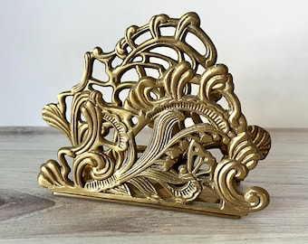 May include: Ornate brass napkin holder with a detailed, openwork design. The holder features swirling patterns and floral motifs, creating an elegant and vintage aesthetic. The brass has a warm, golden hue, adding to its decorative appeal.