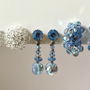 Könnte beinhalten: Drei Paar Ohrclips. Das erste Paar ist weiß und rund. Das zweite Paar ist blau und hat ein Blumenmuster mit einer baumelnden Kristallkugel. Das dritte Paar ist blau und hat einen Kristallhaufen.