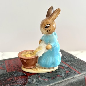 May include: A ceramic figurine of a brown rabbit wearing a light blue dress, pouring from a white cloth into a brown bowl. The figurine is a classic children's character, likely from a storybook.