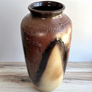 May include: A tall, handcrafted ceramic vase with a wide body and a narrow neck. The vase features a rich, earthy color palette of brown, tan, and black, with a glossy finish. The surface has a textured, organic feel, adding to its unique aesthetic.