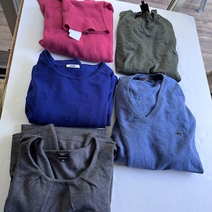 May include: A collection of five long-sleeved sweaters in various colors including pink, olive green, royal blue, light blue, and charcoal gray. The sweaters feature different necklines, including crew necks and v-necks, with some displaying small brand logos.