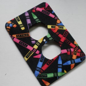 May include: A black electrical outlet cover with a colorful crayon pattern. The crayons are labeled with their colors, including red, orange, yellow, green, blue, and purple.
