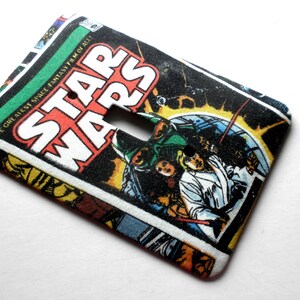 May include: A fabric light switch cover featuring a Star Wars comic book design. The cover has a rectangular shape with a cutout for the switch. The comic book art includes the Star Wars logo and various characters in action.