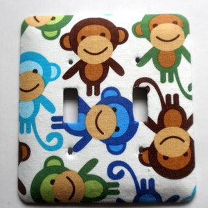 May include: A white light switch cover with a colorful monkey pattern. The monkeys are brown, blue, and green with white backgrounds. The switch cover has two toggle switches.