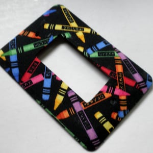 May include: A black light switch cover with a colorful crayon pattern. The crayons are arranged in a diagonal pattern and have the names of the colors written on them.