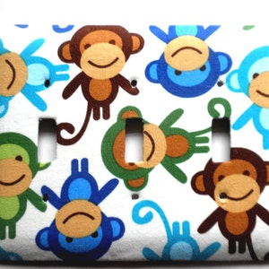 May include: A white light switch cover with a colorful monkey pattern. The monkeys are blue, green, brown, and light blue. The monkeys are smiling and have different expressions.