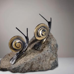May include: Two metal snail sculptures with bronze-colored shells and black bodies are mounted on a gray stone. The snails have long, slender antennae. The shells are intricately detailed with a spiral design. The sculpture is a decorative item.