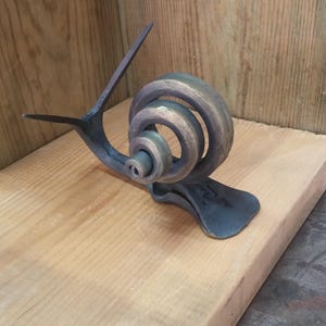 May include: A metal sculpture of a snail with a coiled shell. The snail is made of dark metal with a bronze finish. The sculpture is sitting on a wooden surface.
