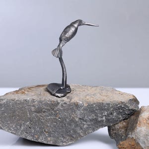May include: A small metal sculpture of a hummingbird perched on a rock. The sculpture is made of steel and has a realistic appearance.