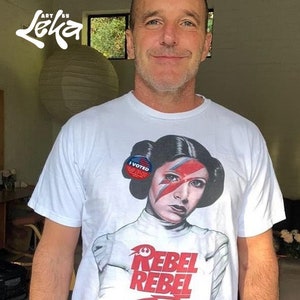 May include: A white t-shirt with a graphic of Princess Leia from Star Wars with a Ziggy Stardust-inspired makeup look. The graphic also includes the text "Rebel Rebel" and a "I Voted" sticker on Leia's ear.