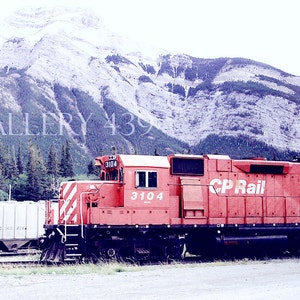 May include: A red CP Rail locomotive with the number 3104 on the side, parked in front of a mountain range.