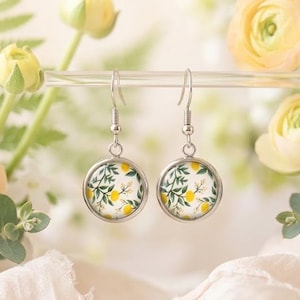 May include: A pair of silver-toned drop earrings featuring circular pendants with a white background and a botanical pattern of yellow lemons and green leaves. The earrings have simple metal hooks for attachment and a polished silver finish.
