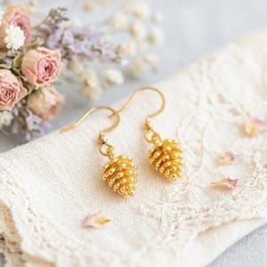Può includere: Orecchini a forma di pigna color oro, con superficie strutturata, presentati su un tessuto di pizzo bianco. Gli orecchini sono posti su uno sfondo di fiori secchi rosa e bianchi, creando un'estetica delicata e naturale.