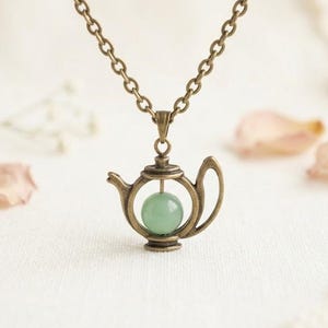 May include: A bronze-colored teapot pendant necklace with a chain. The teapot has a round, light green stone inside. The background is a soft white with a few pink flower petals.