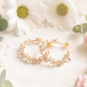 May include: A pair of gold-colored hoop earrings with a delicate leaf design, surrounded by pink and white roses, daisies, and greenery. The earrings are displayed on a white surface with a soft, romantic aesthetic.