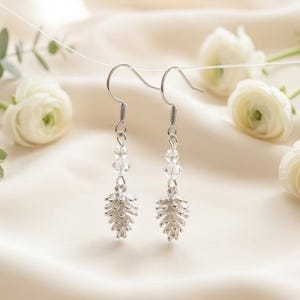 Puede incluir: Pendientes colgantes plateados con abalorios en forma de hoja y cuentas de cristal transparente. Los pendientes cuelgan de ganchos plateados. Flores blancas y vegetación sobre tela color crema.