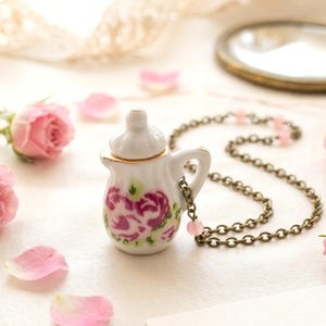 Peut inclure: Un collier pendentif en forme de petite théière en céramique blanche avec un motif floral rose et violet. La chaîne est en bronze, entourée de roses roses, de pétales et d'un miroir vintage. Une carte avec le texte "La Petite Maison" est également présente.
