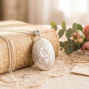 Pode incluir: Colar com medalhão oval prateado com gravura floral, repousando sobre um livro vintage com o texto "POEMS of the HEART". A cena inclui flores cor-de-rosa, óculos e um envelope selado com cera, criando uma estética romântica.