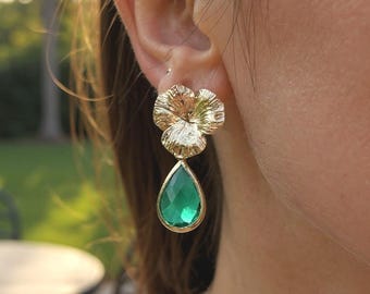 May include: A gold-toned earring featuring a textured, flower-shaped stud at the top and a faceted, teardrop-shaped green gemstone pendant hanging below. The metal has a polished finish, and the green stone is set in a gold-colored frame.