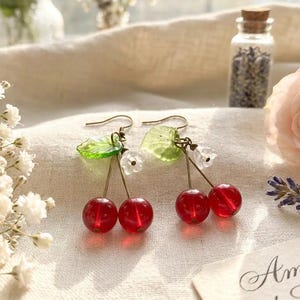 Puede incluir: Un par de pendientes colgantes con dos cuentas esféricas rojas que parecen cerezas, adornadas con pequeñas hojas verdes translúcidas y diminutas cuentas blancas en forma de flor, suspendidas de ganchos de metal dorado sobre una tela.