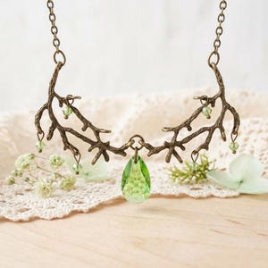 May include: A bronze-toned necklace with a branch-like design. The necklace features small green beads and a teardrop-shaped green pendant. The jewelry is displayed on a lace cloth with small white flowers.