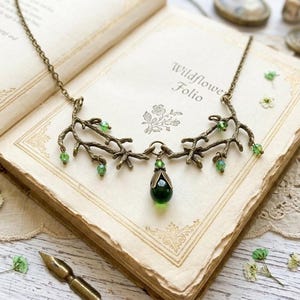 Puede incluir: Un collar de tono bronce con un diseño de ramas adornado con cuentas verdes. El collar descansa sobre un libro abierto titulado "Wildflower Folio". Un colgante en forma de lágrima verde oscuro cuelga del centro. El fondo incluye flores secas y una pluma.