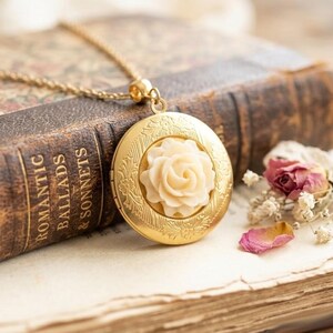 Pode incluir: Um colar dourado com um medalhão com um design de rosa creme, repousando sobre um livro antigo. O livro é marrom com letras douradas que dizem "Romantic Ballads & Sonnets". Pétalas e flores secas estão por perto.