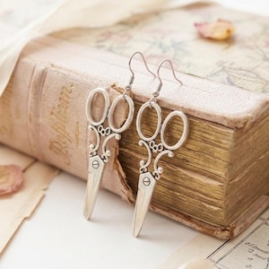 Puede incluir: Un par de pendientes plateados con forma de tijeras y detalles de filigrana intrincados. Los pendientes se exhiben sobre un libro antiguo con una cubierta rosa claro y páginas envejecidas, junto con pétalos de flores secas y una cinta.