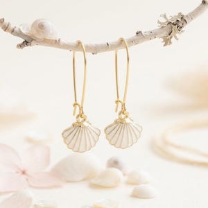 Peut inclure: Une paire de boucles d'oreilles pendantes dorées avec des breloques en forme de coquillage en émail blanc. Les boucles d'oreilles sont dotées de longues tiges dorées fines et sont suspendues à une branche claire, offrant un design épuré et délicat.