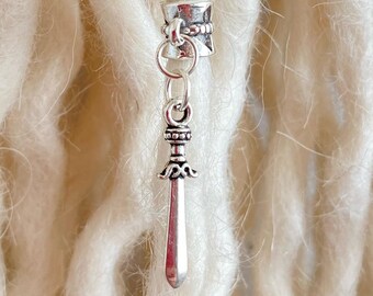 Peut inclure: Une perle de dreadlock argentée avec un charme d'épée. La perle a un design cylindrique avec des détails complexes, et le charme d'épée est suspendu en dessous. Le fond présente plusieurs mèches de dreadlocks blanc cassé.