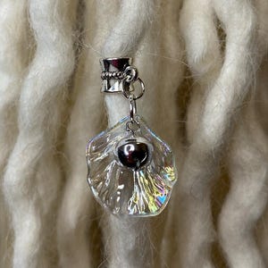 May include: A clear, iridescent dreadlock bead with a silver-colored metal clasp and a small, dark heart-shaped charm. The bead is shaped like a shell and hangs from a silver chain. The background features multiple white dreadlocks.