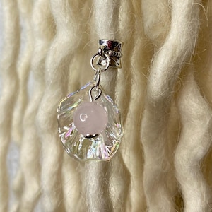Puede incluir: Un dije de cristal transparente y facetado con un centro de piedra rosa, suspendido de una cuenta de metal plateado. El dije cuelga sobre un fondo de rastas gruesas de color crema, con una pared de ladrillos blancos texturizados en el fondo.
