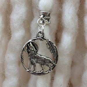 Könnte beinhalten: Eine silberfarbene Dreadlock-Perle mit einem heulenden Wolf-Design in einem kreisförmigen Rahmen. Der Wolf ist detailliert dargestellt, und eine Feder ist ebenfalls enthalten. Die Perle ist an einem silberfarbenen Verschluss befestigt.