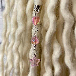 Op de afbeelding: Een zilverkleurig haaraccessoire met een hart, een bol en een sterbedel, allemaal in iriserend roze. Het accessoire is bevestigd aan een bundel off-white, getextureerde hairextensions.