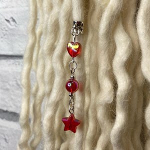 Op de afbeelding: Een zilverkleurige dreadlock-kralenbedel met rode hart-, ronde en stervormige kralen. De bedel is bevestigd aan een gedeelte van dikke, crèmekleurige dreadlocks. De kralen hebben een doorschijnende, iriserende kwaliteit.