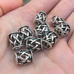 May include: A collection of silver-colored beads with an open, woven design. Each bead is cylindrical with a detailed pattern of intersecting lines, creating a cage-like appearance. The beads are small and appear to be made of metal.