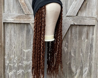 Könnte beinhalten: Eine schwarze Mütze mit langen, braunen Dreadlocks. Die Dreadlocks haben hellbraune und blonde Highlights. Das Haar ist auf einem Schaufensterkopf gestylt, vor einem verwitterten Holzhintergrund.
