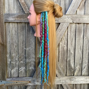 May include: A mannequin head with long blonde hair styled in a bun and cascading, colorful dreadlocks. The dreadlocks are a mix of blue, green, purple, and pink, with silver accents. The background is a weathered wooden door.