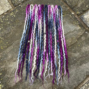 Könnte beinhalten: Eine Sammlung bunter Dreadlocks in Lila-, Blau- und Weißtönen. Die Dreads sind vertikal angeordnet, einige Strähnen sind mit silbernen Akzenten verziert. Die Enden der Dreadlocks haben einen verblassten Ombre-Effekt.
