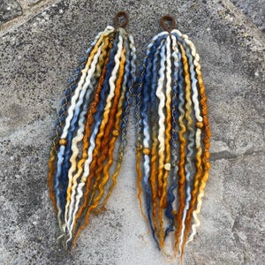 Peut inclure: Deux extensions de dreadlocks faites à la main, dans des tons de bleu, blanc et orange. Chaque dreadlock est orné de fil doré et de perles en bois, et possède une boucle marron en haut pour l'attache. Les dreadlocks sont présentés sur une surface grise texturée.