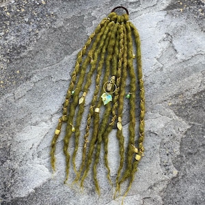 Peut inclure: Dreadlocks vert olive avec des détails dorés et des breloques décoratives. Les dreadlocks sont attachées avec un élastique marron. Les breloques comprennent des perles dorées, des formes de feuilles et un petit pendentif avec une feuille bleu-vert.