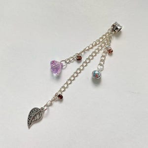 Op de afbeelding: Zilverkleurige bedel met drie bungelende kettingen. De kettingen hebben een gefacetteerde paarse edelsteen, een kleine zilveren bel en een decoratieve bladbedel. Kleine roodbruine kralen zijn langs de kettingen verspreid. De bedel is ontworpen om aan een armband of ketting te worden bevestigd.
