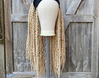 Op de afbeelding: Een pruik met lange, blonde dreadlocks, een zwarte hoofdband en houten kralen, tentoongesteld op een paspop. De paspop is gemonteerd op een grijze standaard. De achtergrond is een verweerde houten deur.