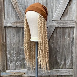May include: A brown turban-style head covering with long, blonde, curly synthetic dreadlocks attached. The wig is displayed on a mannequin head, mounted on a gray stand. The background is a weathered wooden door.